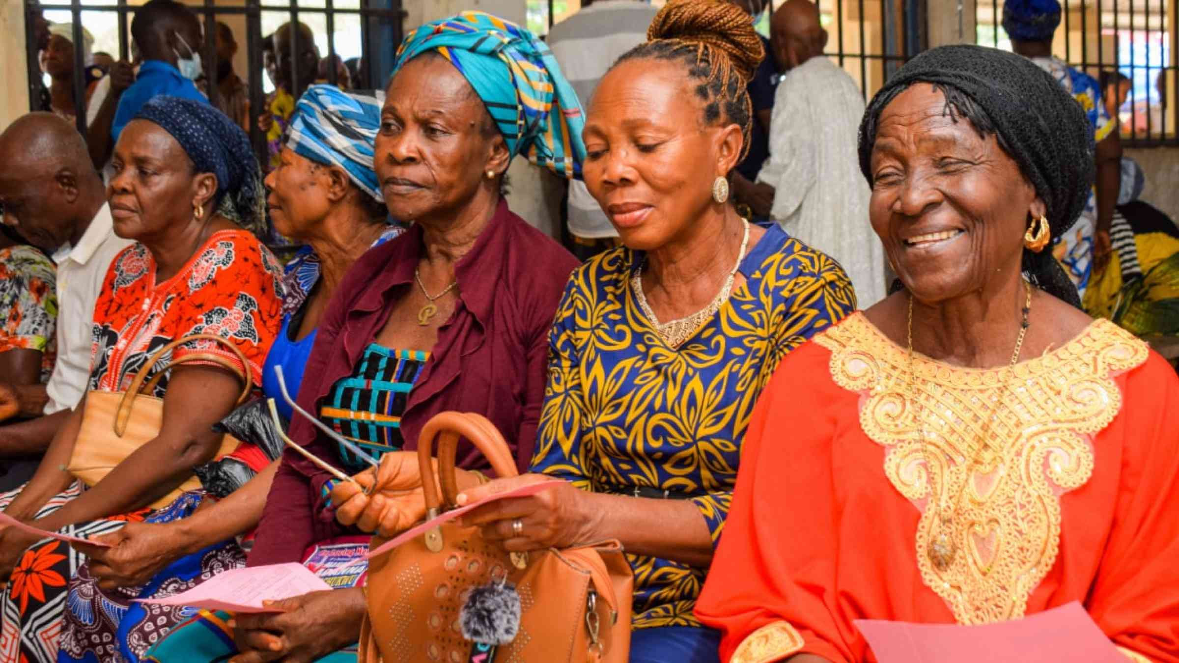 Four African women sitting next to each other on a bench in a room of people.